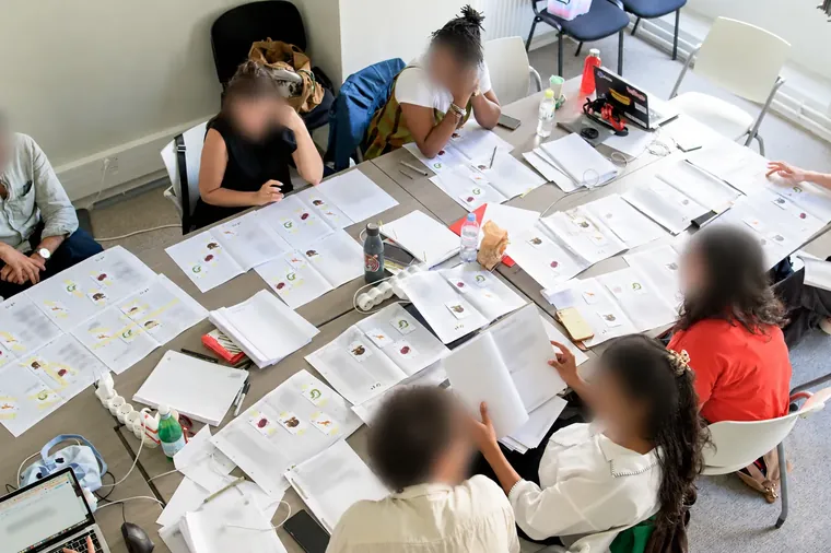 Vue plongeante sur une table de travail couverte de carnets et de cartes, autour de laquelle sont assis des participant·es aux visages floutés.